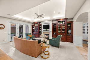 Living area with arched walkways, a glass covered fireplace, a tray ceiling, and a ceiling fan