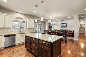 Kitchen with dark brown cabinets, light stone counters, tasteful backsplash, stainless steel dishwasher, and decorative light fixtures