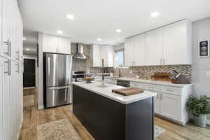 Kitchen featuring appliances with stainless steel finishes, white cabinetry, light wood-style floors, wall chimney exhaust hood, and a center island