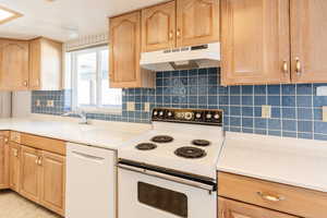 Kitchen with white appliances, light brown cabinetry, under cabinet range hood, and backsplash