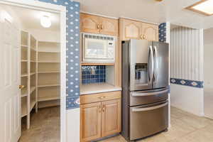 Kitchen with wallpapered walls, stainless steel refrigerator with ice dispenser, white microwave, light brown cabinets, and light countertops