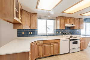 Kitchen with white appliances, glass insert cabinets, backsplash, under cabinet range hood, and light stone countertops