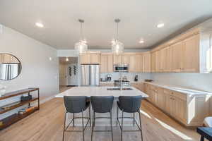 Kitchen with light brown cabinets, stainless steel appliances, pendant lighting, a kitchen bar, and an island with sink