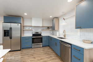 Kitchen featuring blue cabinetry, appliances with stainless steel finishes, light wood-type flooring, light stone countertops, and open shelves