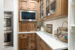 Kitchen featuring stainless steel double oven, brown cabinets, built in microwave, light countertops, and glass insert cabinets