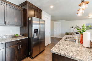 Kitchen featuring dark brown cabinetry, stainless steel fridge, light stone countertops, light wood-style floors, and hanging light fixtures