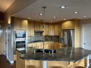 Kitchen with stainless steel appliances, dark stone countertops, decorative light fixtures, an island with sink, and a breakfast bar area