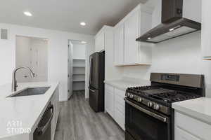 Kitchen with appliances with stainless steel finishes, wall chimney range hood, light stone countertops, white cabinetry, and light wood finished floors