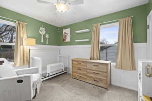 Carpeted bedroom with a wainscoted wall, a decorative wall, radiator heating unit, multiple windows, and a ceiling fan