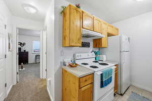 Kitchen with white electric stove, light countertops, under cabinet range hood, light colored carpet, and light brown cabinetry