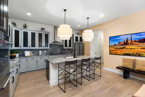 Kitchen featuring decorative light fixtures, a breakfast bar, a center island with sink, light stone counters, and wall chimney exhaust hood