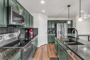 Kitchen with green cabinets, stainless steel appliances, decorative light fixtures, light wood finished floors, and dark stone countertops