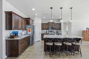 Kitchen with dark brown cabinets, hanging light fixtures, a kitchen bar, a center island with sink, and light stone counters