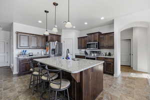 Kitchen featuring dark brown cabinets, arched walkways, appliances with stainless steel finishes, hanging light fixtures, and a center island with sink