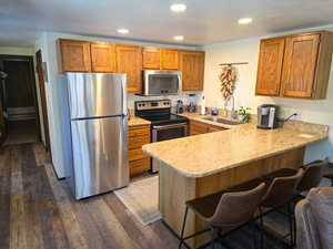 Kitchen with appliances with stainless steel finishes, light stone counters, a peninsula, dark wood-style flooring, and a breakfast bar area
