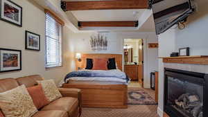 Bedroom featuring a glass covered fireplace, beamed ceiling, ensuite bath, and light carpet