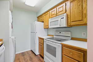 Kitchen with white appliances, light countertops, dark wood-style flooring, stacked washer / drying machine, and brown cabinets