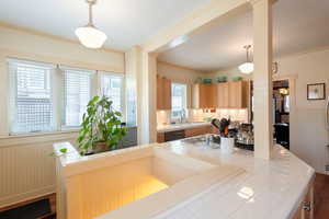 Kitchen with tile countertops, a wainscoted wall, light brown cabinetry, decorative light fixtures, and ornamental molding