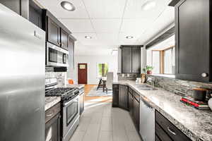 Kitchen with appliances with stainless steel finishes, a paneled ceiling, recessed lighting, light stone counters, and backsplash