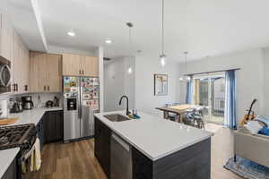 Kitchen with stainless steel appliances, hanging light fixtures, an island with sink, dark wood-style flooring, and light brown cabinetry