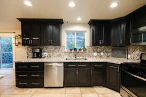 Kitchen featuring dark cabinets, appliances with stainless steel finishes, light stone countertops, decorative backsplash, and recessed lighting