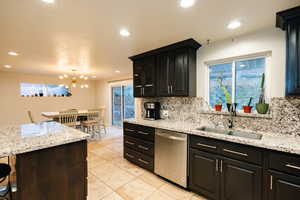 Kitchen featuring dark cabinets, light stone counters, dishwasher, backsplash, and a chandelier