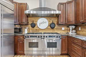 Kitchen featuring wall chimney range hood, high quality appliances, and dark stone counters