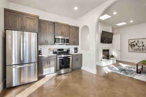 Kitchen with appliances with stainless steel finishes, finished concrete flooring, tasteful backsplash, dark brown cabinetry, and a glass covered fireplace