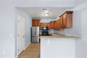 Kitchen featuring black appliances, brown cabinetry, light wood finished floors, a peninsula, and light stone counters