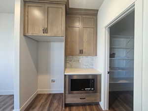 Kitchen with decorative backsplash, stainless steel microwave, dark wood-style flooring, light stone countertops, and brown cabinetry