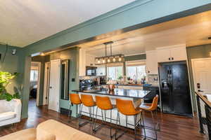 Kitchen featuring a breakfast bar area, black appliances, white cabinets, decorative light fixtures, and dark wood finished floors