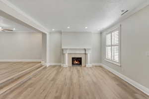 Unfurnished living room with a warm lit fireplace, light wood finished floors, wide-louvered shutters, recessed lighting, and crown molding