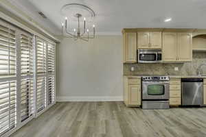 Kitchen featuring light brown cabinets, appliances with stainless steel finishes, hanging light fixtures, decorative backsplash, and ornamental molding