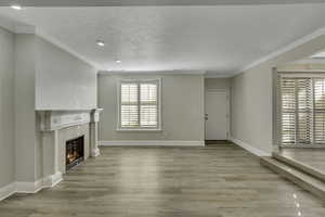 Unfurnished living room with a textured ceiling, a fireplace, light wood finished floors, crown molding, wide-louvered shutters, and recessed lighting