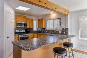 Kitchen with dark countertops, appliances with stainless steel finishes, backsplash, light tile patterned flooring, and beam ceiling