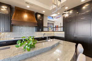 Kitchen featuring light stone countertops, custom exhaust hood, glass insert cabinets, decorative light fixtures, and backsplash