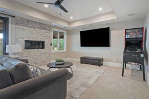 Carpeted living room with ceiling fan, a raised ceiling, a stone fireplace, and recessed lighting