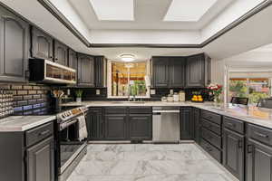 Kitchen featuring stainless steel appliances, tasteful backsplash, light marble finish flooring, light stone countertops, and a tray ceiling