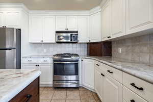 Kitchen with stainless steel appliances, light tile patterned floors, decorative backsplash, light stone countertops, and dual tone cabinetry