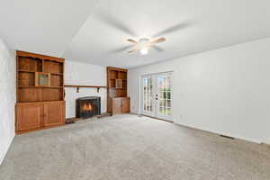 Unfurnished living room featuring a warm lit fireplace, light colored carpet, ceiling fan, built in features, and french doors
