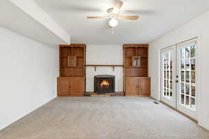 Unfurnished living room featuring a warm lit fireplace, light colored carpet, a ceiling fan, and french doors