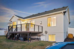 Back of house at dusk featuring a shingled roof, a deck, stucco siding, a yard, and stairway