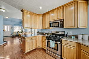 Kitchen with appliances with stainless steel finishes, a textured ceiling, a peninsula, light wood finished floors, and light brown cabinetry