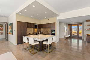 Kitchen featuring a kitchen bar, dark brown cabinets, a center island with sink, decorative backsplash, and modern cabinets