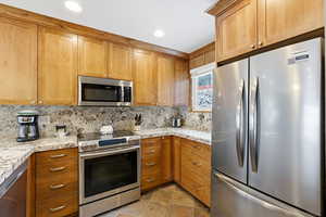 Kitchen with appliances with stainless steel finishes, decorative backsplash, brown cabinetry, light stone counters, and recessed lighting