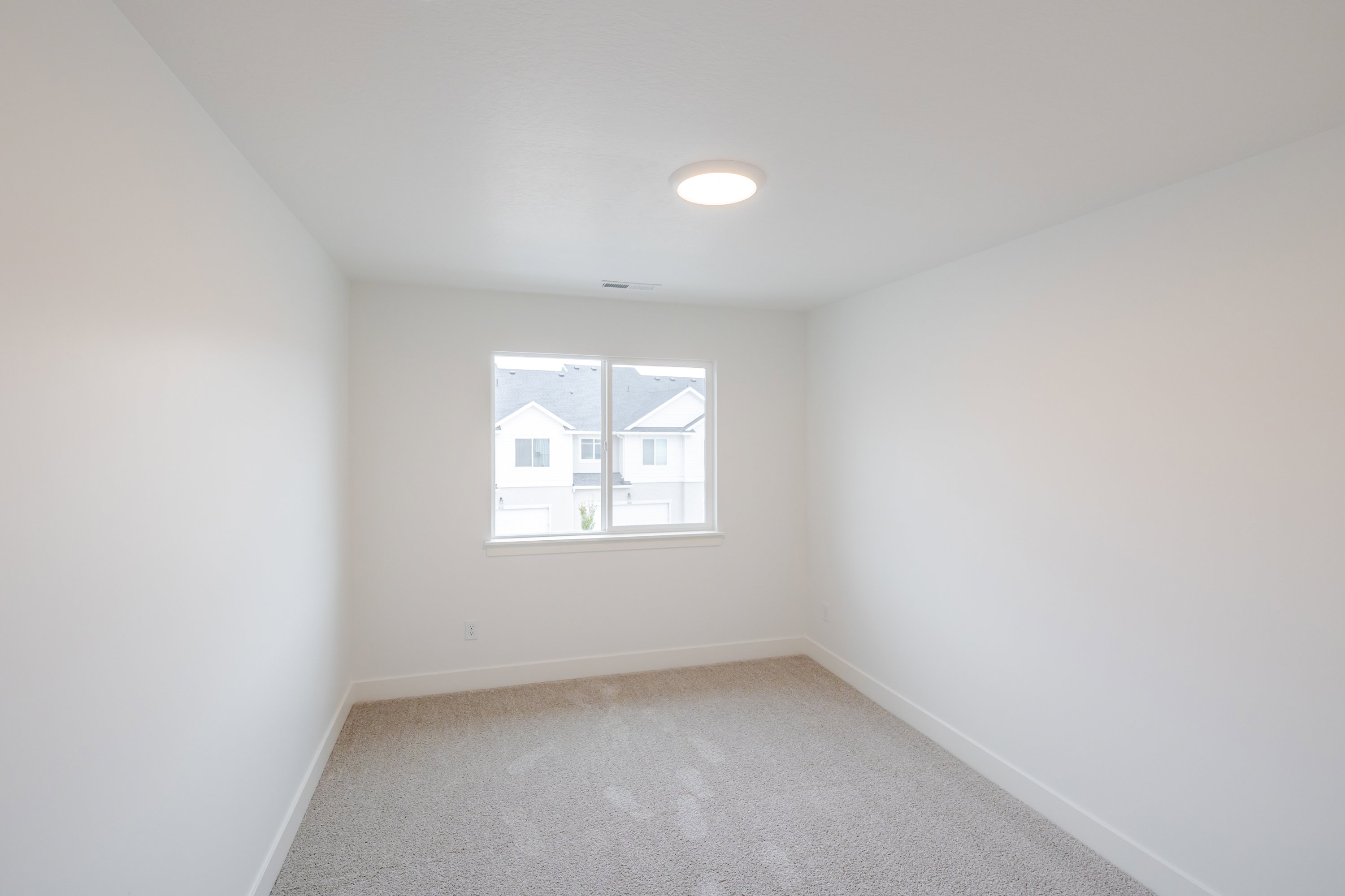 Light-filled interior space featuring a large double window, neutral carpeting, white baseboards, a flush-mount ceiling light fixture, and crisp white wall finishes