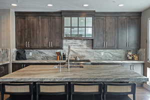 Kitchen with a breakfast bar, backsplash, light stone countertops, dark brown cabinets, and recessed lighting
