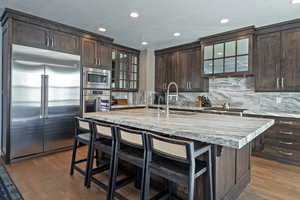 Kitchen featuring dark brown cabinets, built in appliances, light stone counters, a breakfast bar, and backsplash