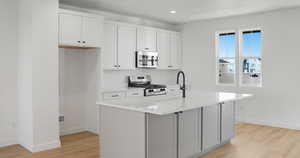 Kitchen with white cabinets, stainless steel appliances, a center island with sink, light wood-type flooring, and light stone counters