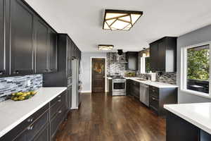 Kitchen featuring decorative backsplash, light stone countertops, dark wood-type flooring, appliances with stainless steel finishes, and dark cabinetry